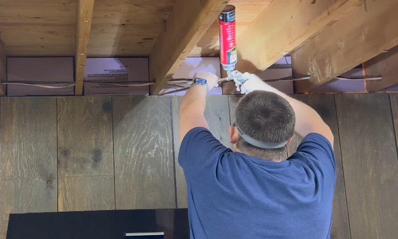 Man in blue shirt sealing gap between ceiling joists with red spray foam can